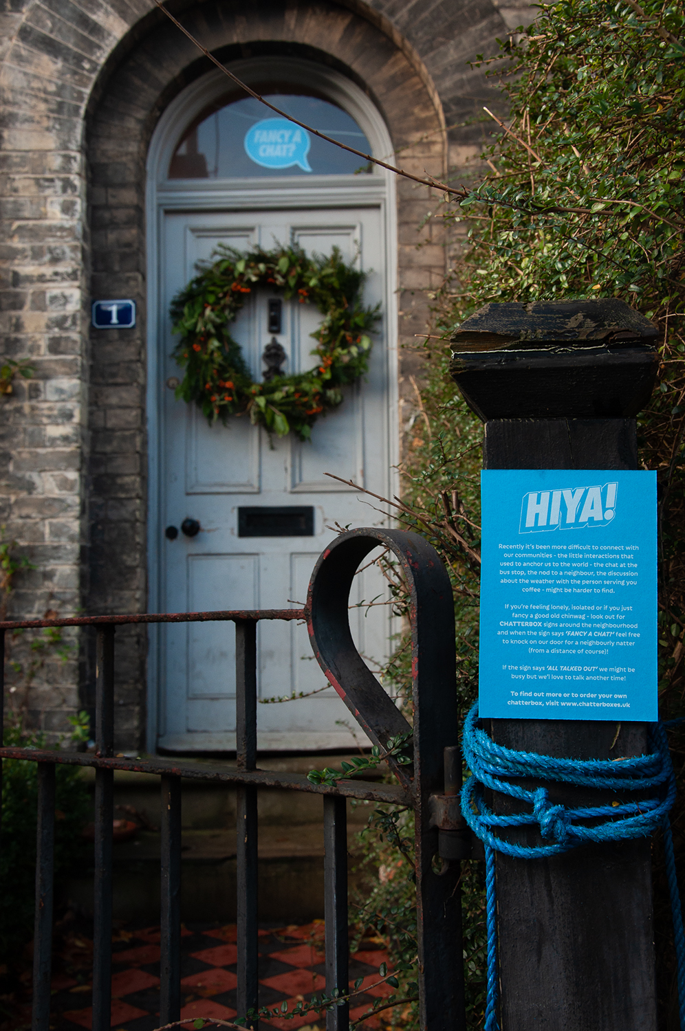 A bright blue sign with the heading HIYA is attached to a gatepost. There is further text explaining the Chatterbox project. In the background, a blue speech bubble shaped sign can be seen hanging in the transom window above the front door.