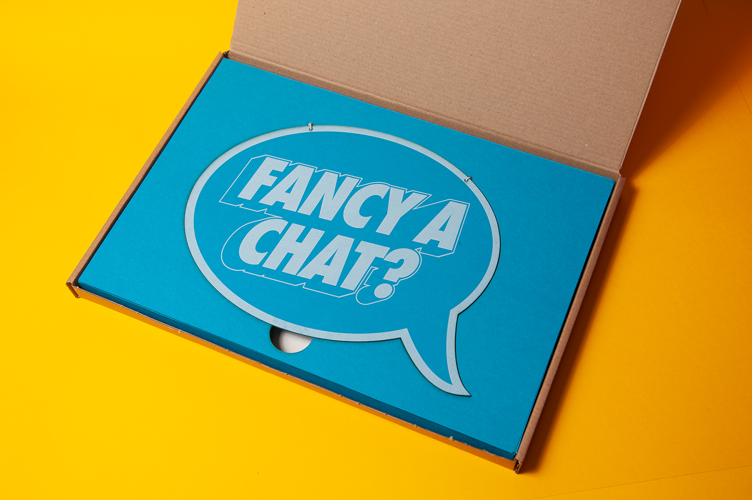 A photograph of the cardboard box, open, on a yellow background. Inside the box is a bright blue insert which holds a bright blue speech bubble shaped sign.