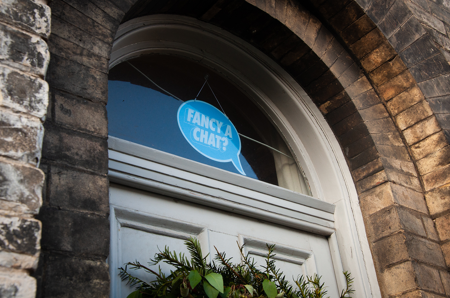 A photograph of a bright blue speech bubble shaped sign in a transom window above a front door. The text in the speech bubble reads Fancy a Chat?